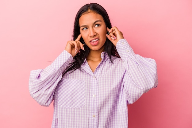 Young latin woman isolated on pink background  covering ears with fingers, stressed and desperate by a loudly ambient.