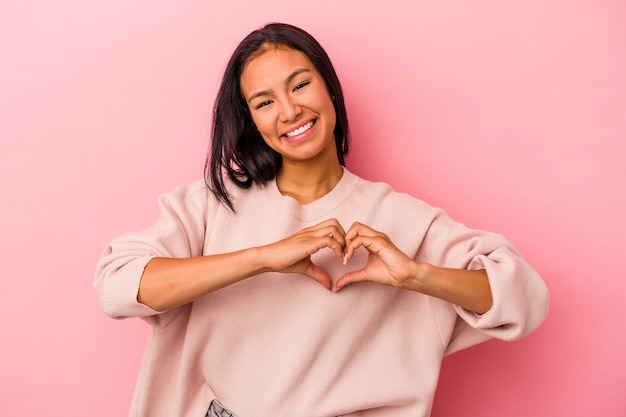 Young latin woman isolated on pink background  smiling and showing a heart shape with hands.