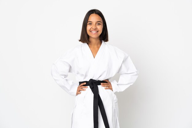 Young latin woman isolated on white wall doing karate posing with arms at hip and smiling