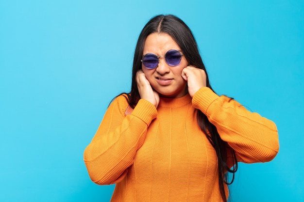 Young latin woman looking angry, stressed and annoyed, covering both ears to a deafening noise, sound or loud music