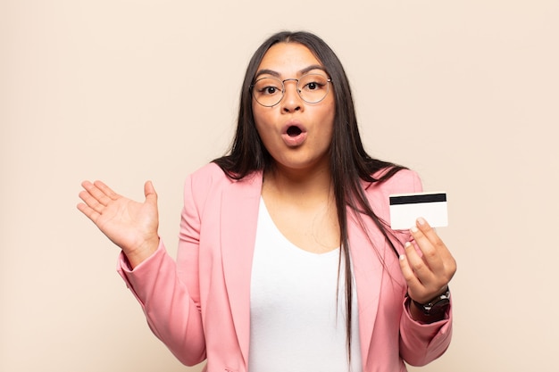 Young latin woman looking surprised and shocked, with jaw dropped holding an object with an open hand on the side