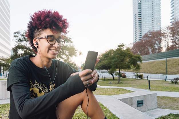 Young latin woman outdoors at sunset sitting amused checking phone smiling copy space