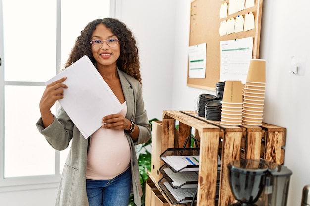 Young latin woman pregnant smiling confident holding document at office