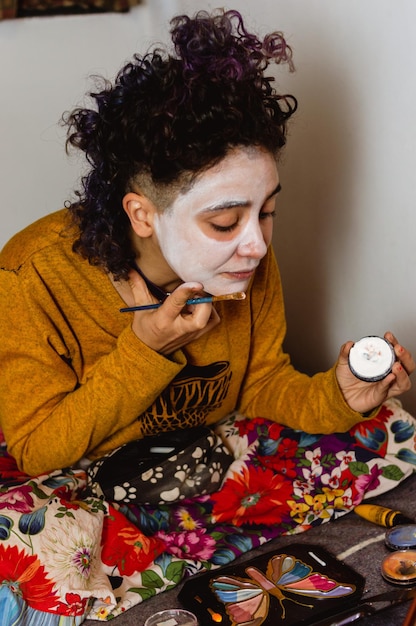 Young latin woman sitting on her bed applying catrina makeup with a brush and white paint