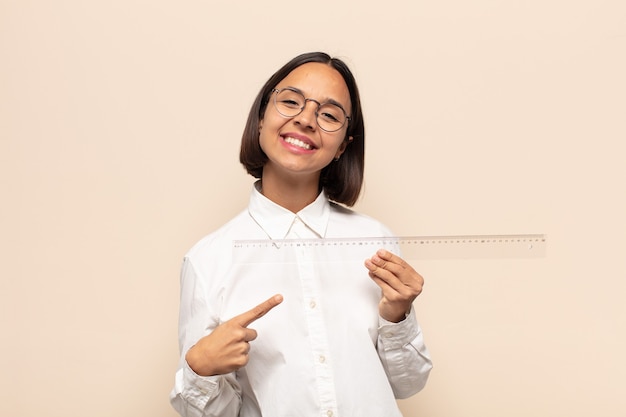 Young latin woman smiling cheerfully, feeling happy and pointing to the side and upwards, showing object in copy space