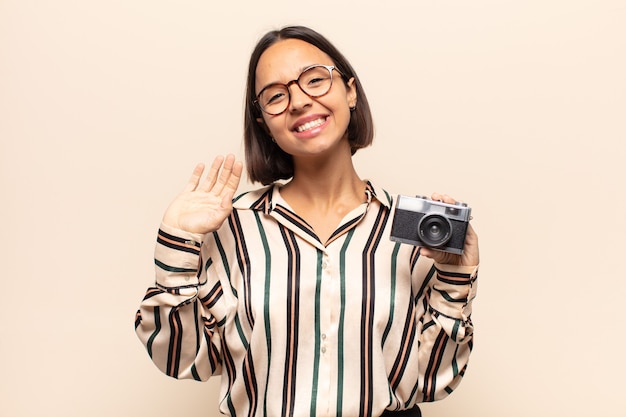 Young latin woman smiling happily and cheerfully, waving hand, welcoming and greeting you, or saying goodbye