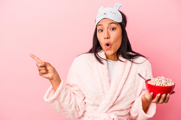 Young latin woman wearing pajama holding a bowl of cereals isolated on pink background  pointing to the side