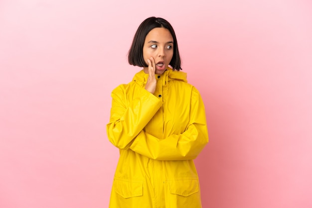 Young latin woman wearing a rainproof coat over isolated background whispering something with surprise gesture while looking to the side