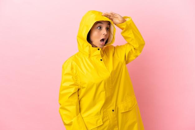 Young latin woman wearing a rainproof coat over isolated background with surprise expression while looking side