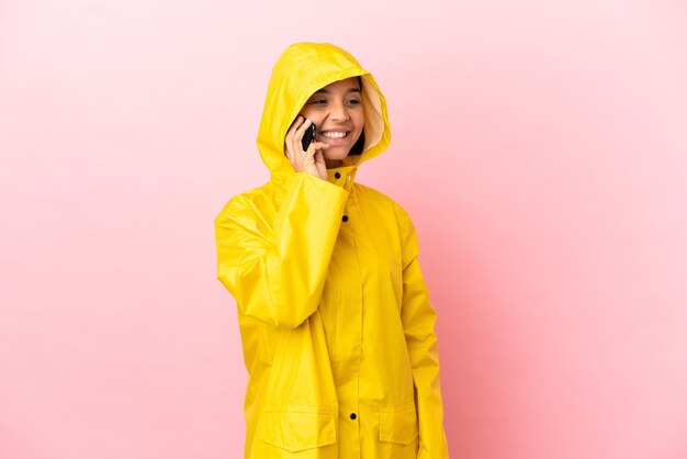 Young latin woman wearing a rainproof coat over isolated wall keeping a conversation with the mobile phone