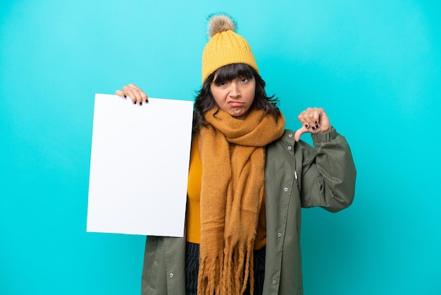 Young latin woman wearing winter jacket isolated on blue background holding an empty placard and doing bad signal