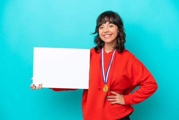 Young latin woman with medals isolated on blue background holding an empty placard with happy expression