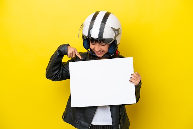 Young latin woman with a motorcycle helmet isolated on yellow background holding an empty placard with happy expression and pointing it