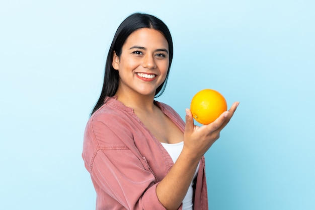 Young latin woman woman isolated on blue holding an orange