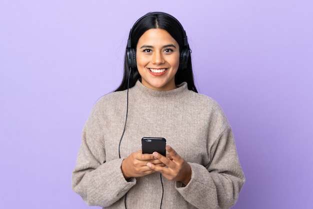 Young latin woman woman on purple wall listening music with a mobile and looking front