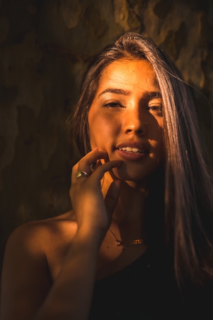 A young Latina brunette leaning against a tree with a slight light on her face. Portrait with smiling