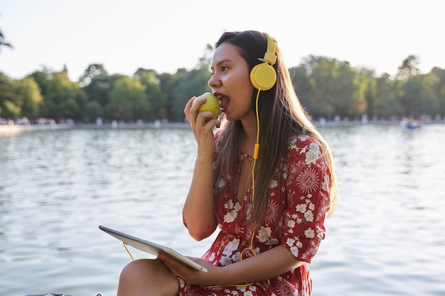 Young latina sitting in a park with a tablet and headphones on bites into an apple