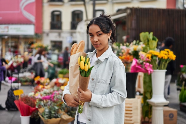 Young Latina woman smiling while buying yellow tulips and bread from a street vendor's stall