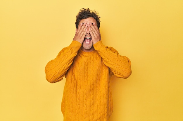 Young Latino man posing on yellow background covers eyes with hands smiles broadly waiting