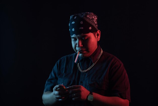 Young Latino man in urban clothes style with a cigarette in his mouth about to light it, illuminated by colored lights on a black background