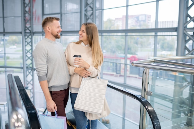 Young laughing dates looking at one another while standing on escalator, enjoying shopping in the mall and talking