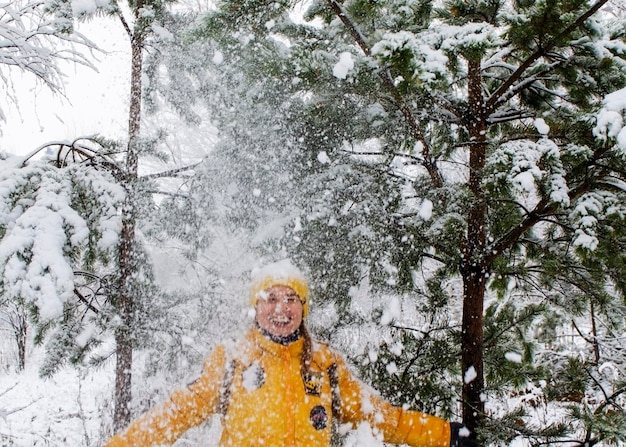 Young laughing happy Caucasian woman in yellow jacket under falling snow from pine  in winter funnny