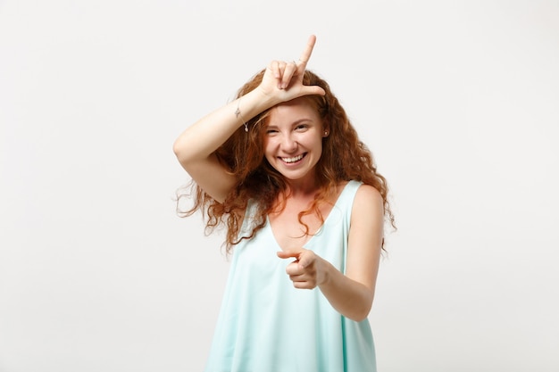 Young laughing redhead woman in casual light clothes posing isolated on white wall background. People lifestyle concept. Mock up copy space. Showing loser gesture, pointing index finger on camera.