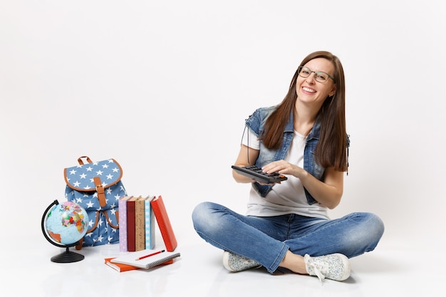 Young laughing smart woman student holding and using calculator solving math equations sitting near globe, backpack, school books isolated