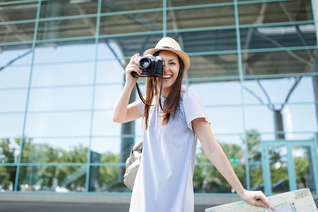 Young laughing traveler tourist woman take pictures on retro vintage photo camera holding paper map at international airport