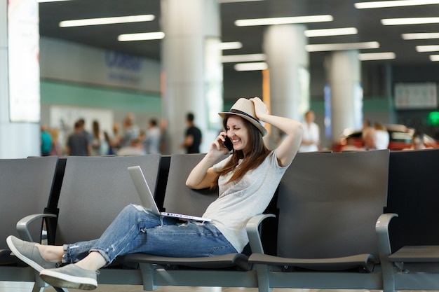 Young laughing traveler tourist woman working on laptop, talk on mobile phone, calls friend at airport