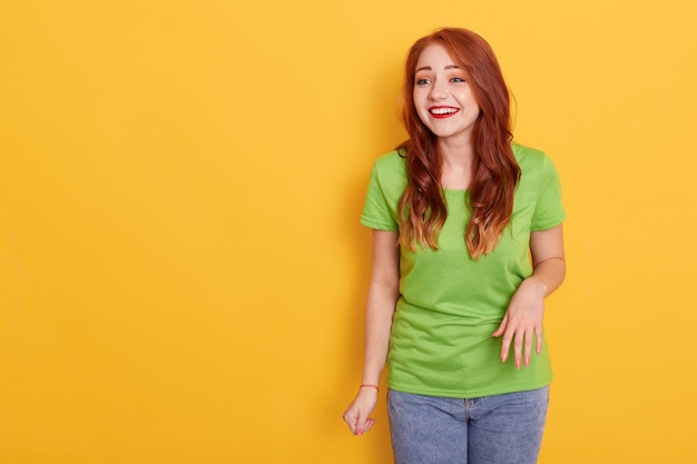 Young laughing woman against yellow background, wearing casual green t shirt and jeans, looking aside, copy space for advertisement or promotional text.