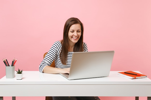 Young laughing woman in casual clothes working on project with laptop while sitting at office isolated on pastel pink background. Achievement business career concept. Copy space for advertisement.