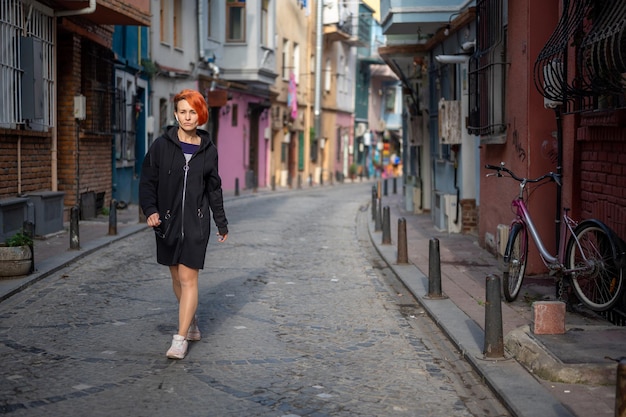 Young lesbian woman confidently walks down the street and looks ahead. Young cheerful woman with bright red hair walks down a narrow street. attractive Young woman walks along narrow city street