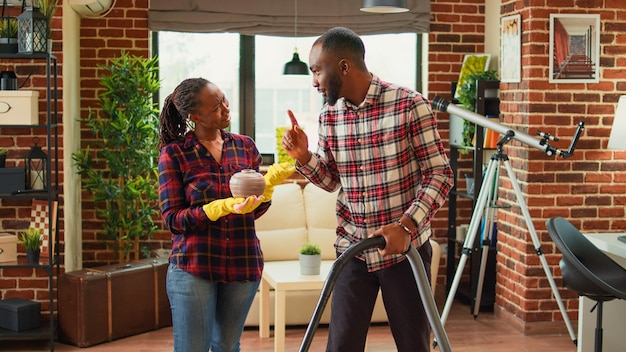 Young life partners doing household chores and cleaning apartment with vacuum cleaner. African american woman sweeping dust off of furniture with rags and washing solution, furniture polish.