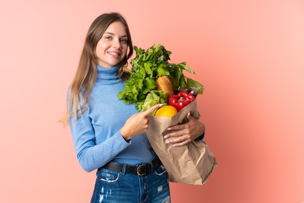 Young Lithuanian woman holding a grocery shopping bag pointing finger to the side