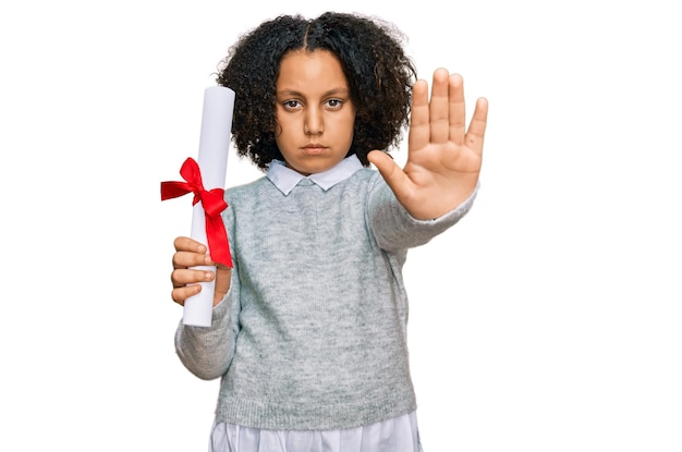 Young little girl with afro hair holding graduate degree diploma with open hand doing stop sign with serious and confident expression defense gesture