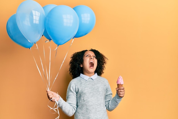 Young little girl with afro hair holding ice cream and blue balloons angry and mad screaming frustrated and furious, shouting with anger looking up.