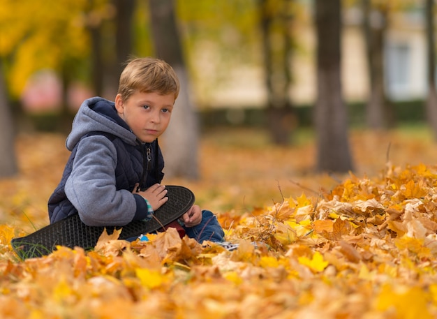 A young, lonely boy with his skateboard sitting in brown fall leaves at a park.