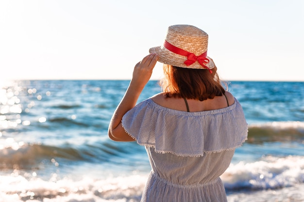 Young lonely caucasian redhaired girl in a straw hat and a summer dress with her back on the beach near the sea and looks