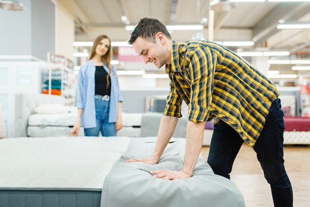 Young love couple checks mattress in furniture store showroom. Man and woman looking samples for bedroom in shop, husband and wife buys goods for modern home interior