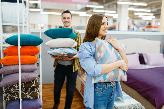 Young love couple holds pillows in furniture store. Man and woman looking samples for bedroom in shop, husband and wife buys goods for modern home interior