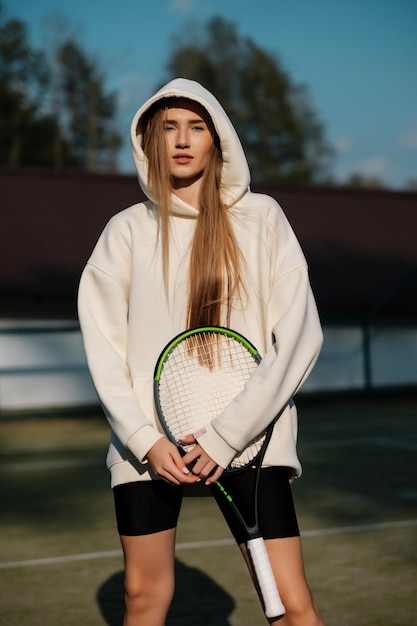 Young lovely long hair girl athlete in white hoodie and tight black shorts with tennis racket outdoors