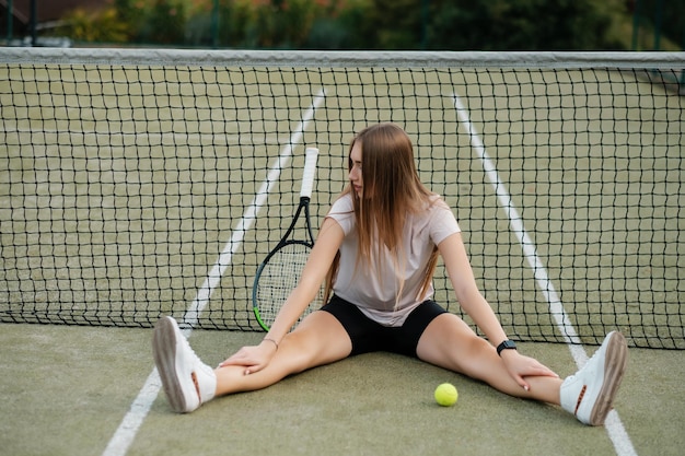 Young lovely long hair girl athlete in white Tshirt and tight black shorts with tennis racket outdoors Female in sport cap sitting at the tennis net