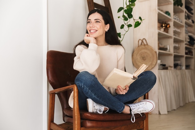 young lovely woman resting in apartment and reading book while sitting on chair