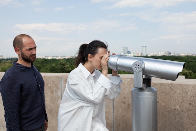 Young lovers spending relationship anniversary on building terrace enjoying summer holiday. Woman looking through telescope at metropolitan city admiring beautiful landscape from observation point