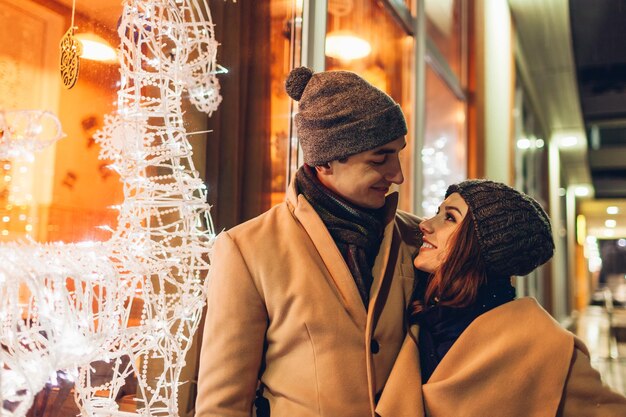 Young loving couple hugging by holiday cafe showcase with decorated reindeers at night