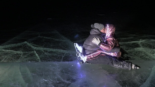 Young loving couple skating at ice rink at night Man and woman kiss hug have fun learn to skate Making love on ice Beautiful clear ice with cracks on lake Ice illuminated backlit from below