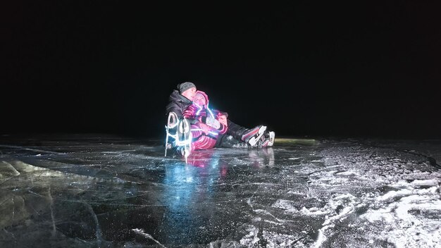 Young loving couple skating at ice rink at night Man and woman kiss hug have fun learn to skate Making love on ice Beautiful clear ice with cracks on lake Ice illuminated backlit from below