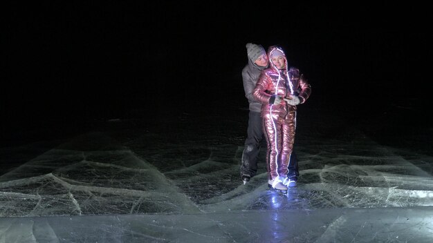 Young loving couple skating at ice rink at night Man and woman kiss hug have fun learn to skate Making love on ice Ice illuminated backlit below