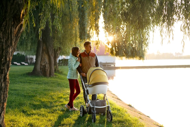 A young loving family walks by the lake with a stroller smiling parents couple with baby pram in